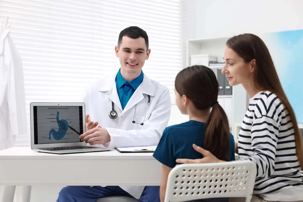 Madre e hija sentadas en un consultorio frente al gastroenterólogo pediatra que explica algo en una imagen del monitor.
